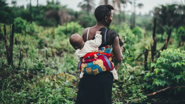 A woman picking plants carries an infant on her back.